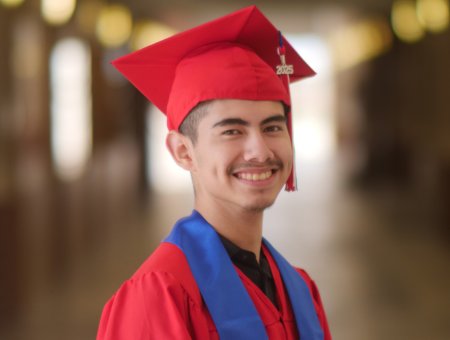 Joab in a red graduation cap and gown with a blue stole, smiling in a hallway.