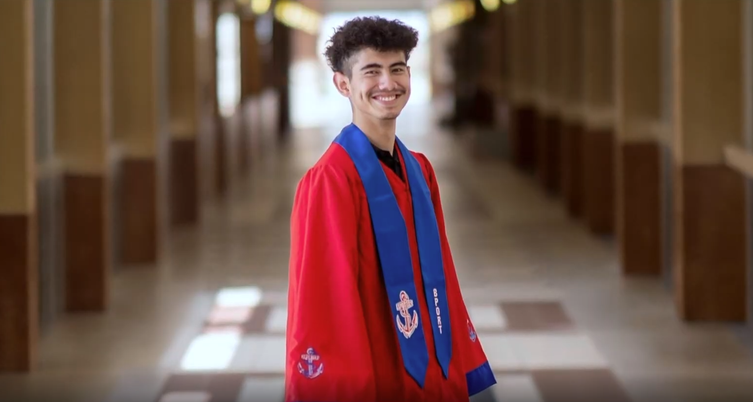 Joab in a red graduation cap and gown with a blue stole, smiling in a hallway.